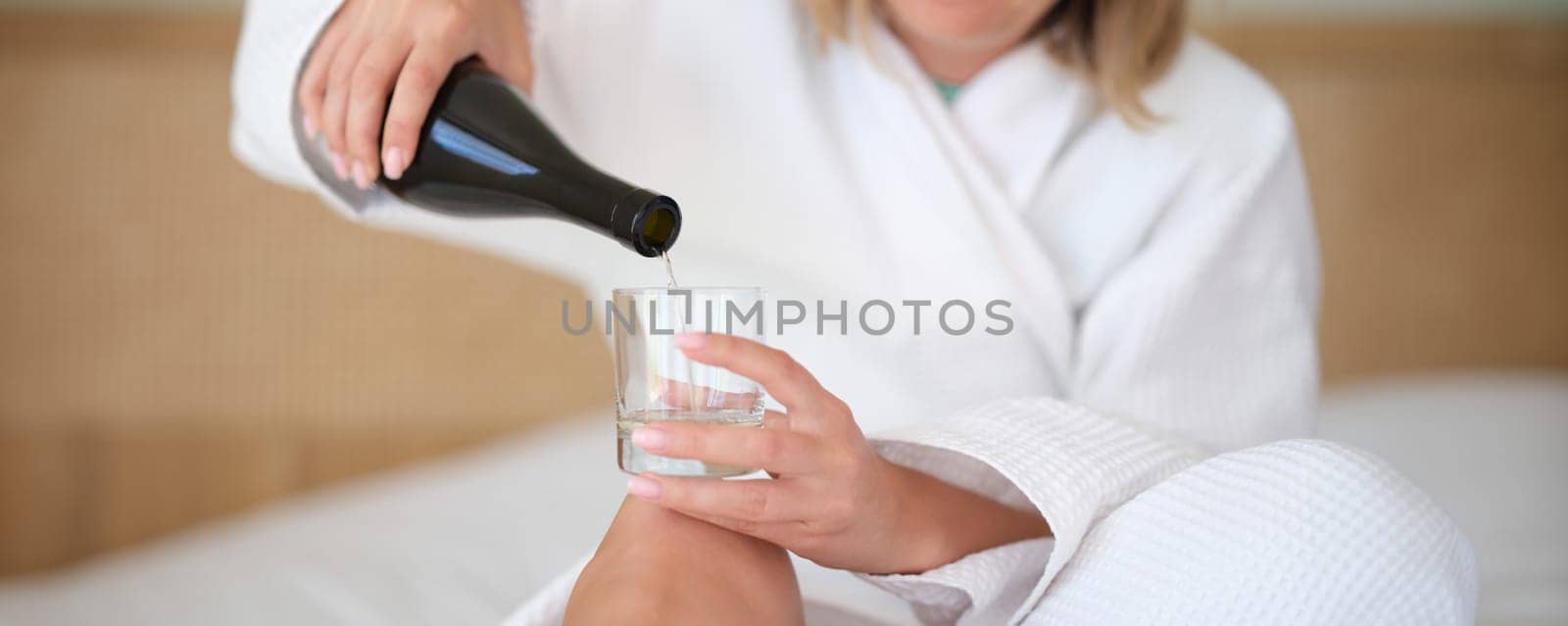Woman in bathrobe on bed pours water or white wine into glass alone by kuprevich