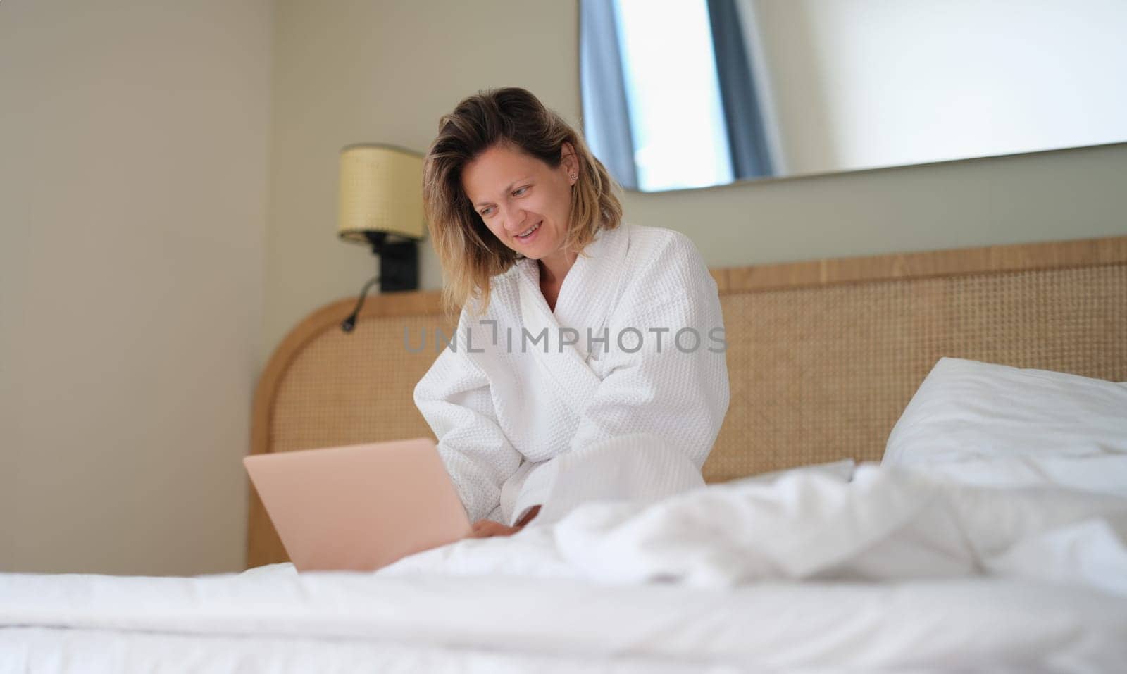 Woman sits at home on bed in white bathrobe and works on laptop. Work from home concept of quarantine and remote work freelancing