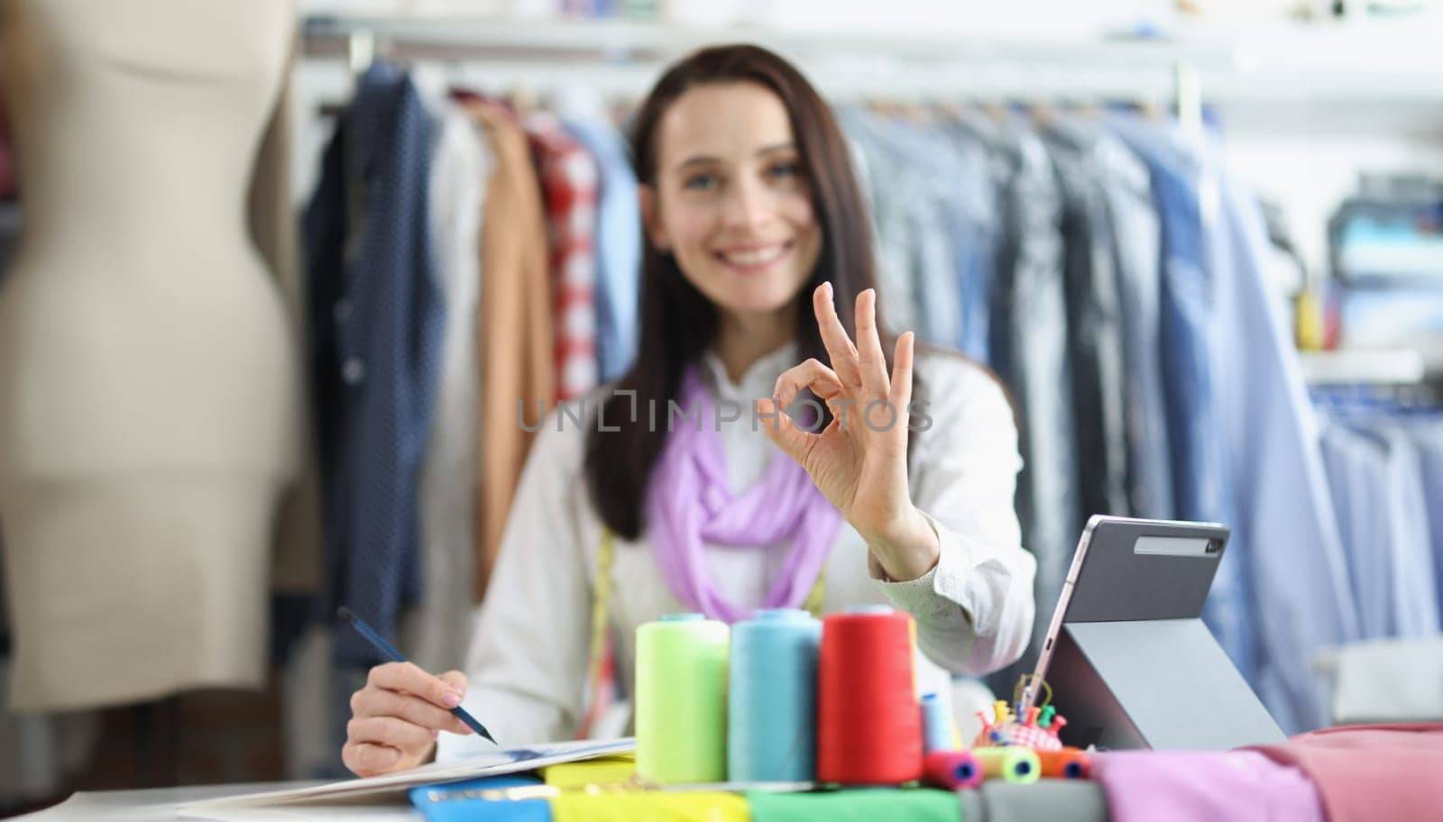 Dressmaker woman holding ok gesture while working in studio. Recommendations for tailoring stylish fashionable clothes and a successful atelier