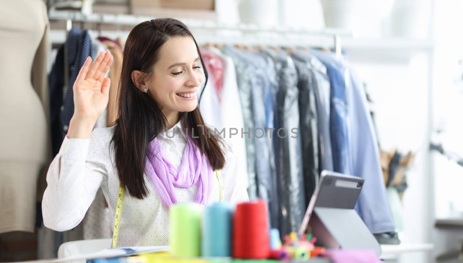 Smiling female designer or seamstress talking on webcam while giving online consultation to client. Dressmaker sits at workplace in front of tablet