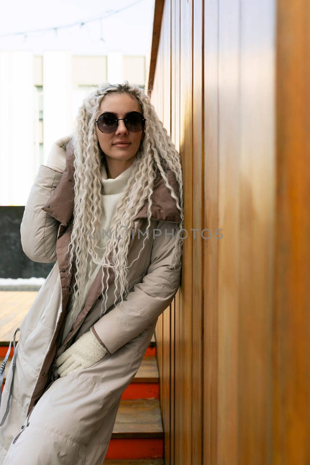 A woman is standing against a wall, wearing a coat and sunglasses. She appears relaxed, with one arm casually draped against the wall. The sunglasses add a touch of mystery to her demeanor.