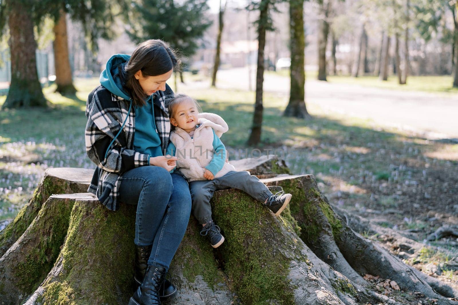 Little smiling girl sits on a huge stump leaning against her mother in the spring forest. High quality photo