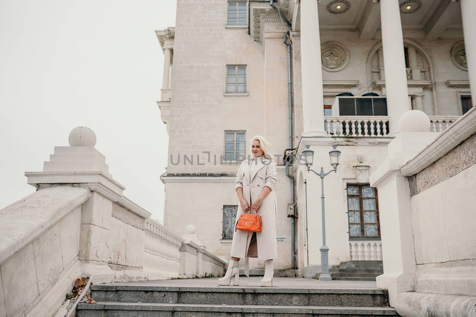 A woman wearing a white coat and holding an orange purse stands on a set of stairs in front of a large white building. by Matiunina
