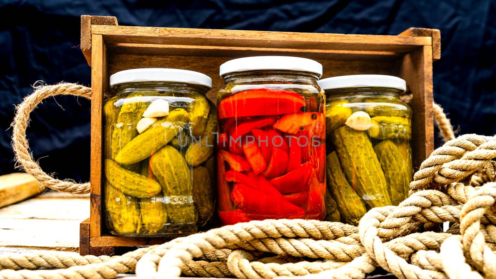 Glass jars with pickled red bell peppers and pickled cucumbers (pickles) isolated in wooden crate. Jars with variety of pickled vegetables. Preserved food concept in a rustic composition. by vladispas
