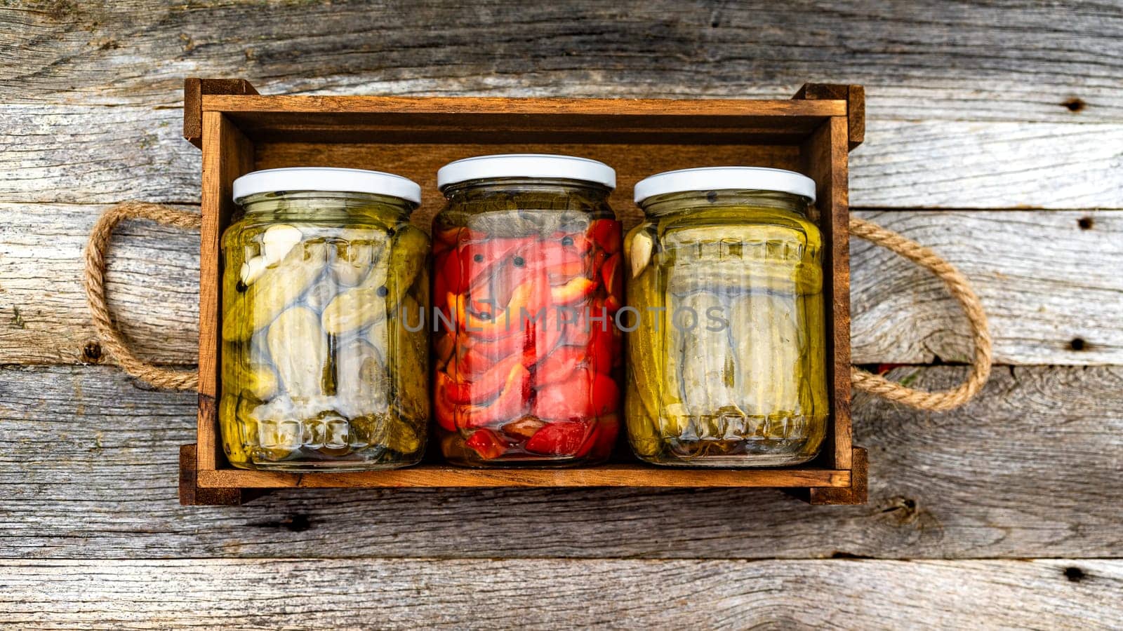 Glass jars with pickled red bell peppers and pickled cucumbers (pickles) isolated in wooden crate. Jars with variety of pickled vegetables. Preserved food concept in a rustic composition.