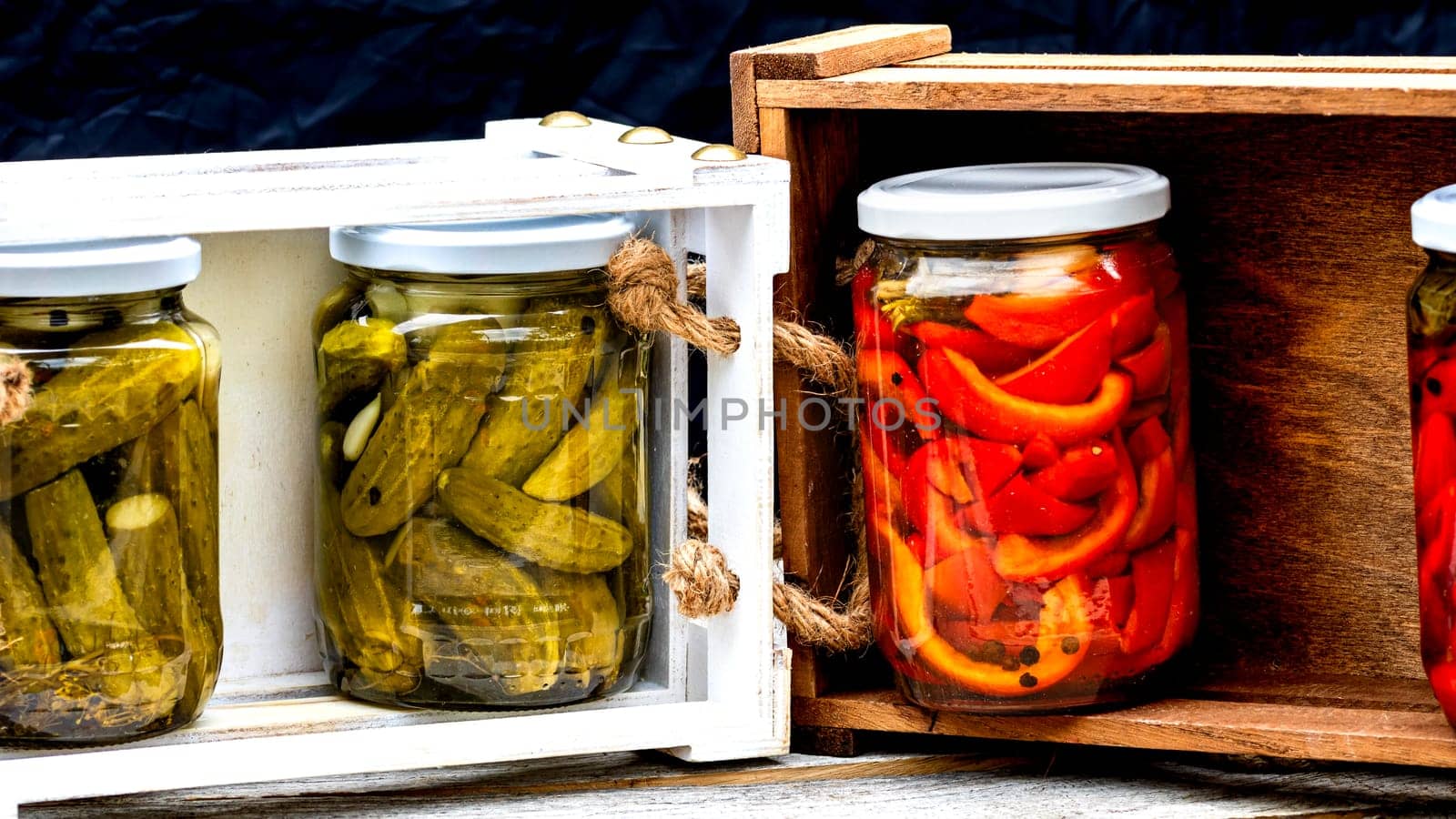 Wooden crate with glass jars with pickled red bell peppers and pickled cucumbers (pickles) isolated. Jars with variety of pickled vegetables. Preserved food concept in a rustic composition. by vladispas