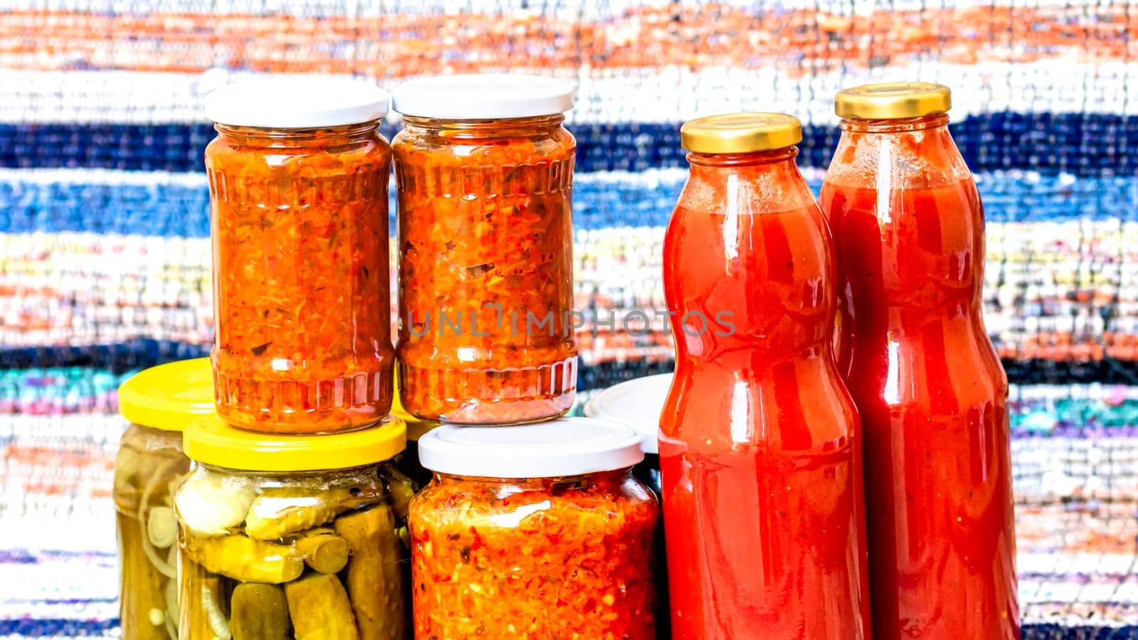 Glass jars with pickled red bell peppers and pickled cucumbers (pickles) isolated. Jars with variety of pickled vegetables, jars with zacusca and bottles with tomatoes sauce. Preserved food concept.