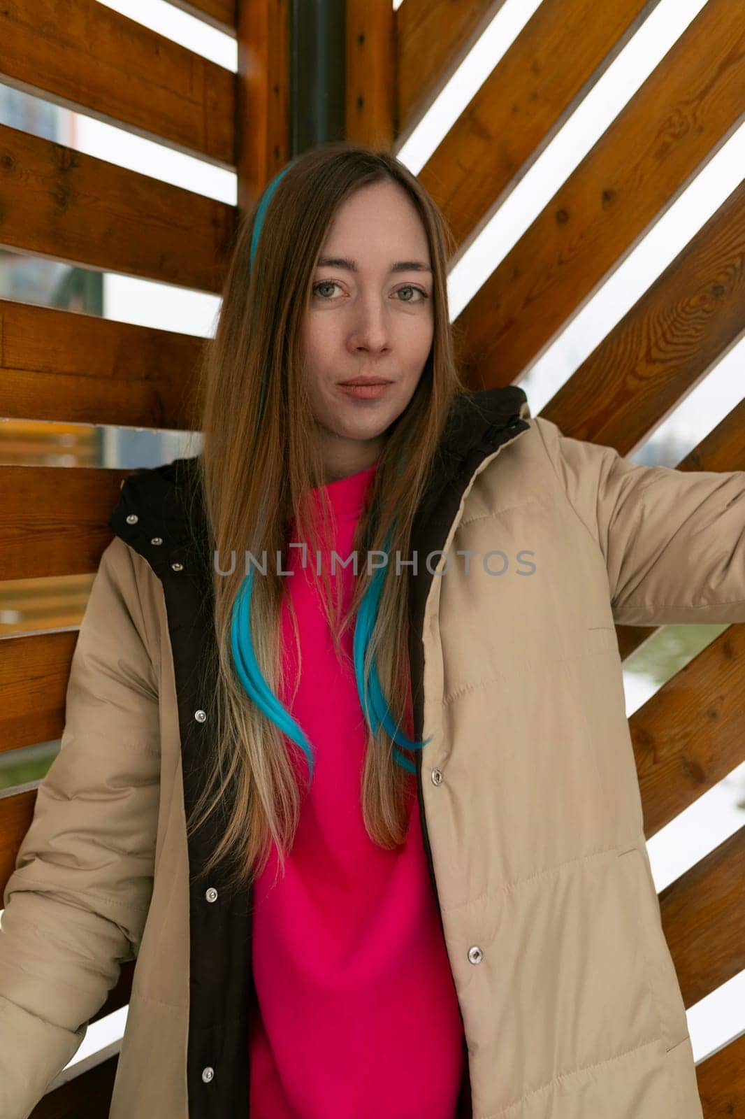 A woman standing upright in front of a weathered wooden wall. She is gazing straight ahead, with a neutral expression on her face. The wooden planks are aged and textured, adding a rustic element to the scene.