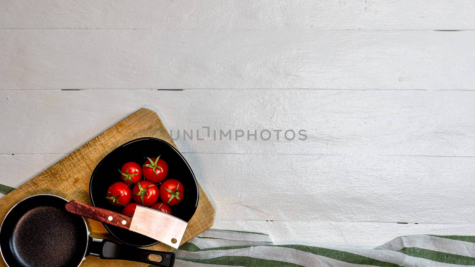 Top view of knife, small pan and fresh ripe cherry tomatoes in small black bowl on a rustic white wooden table. Ingredients and food concept by vladispas