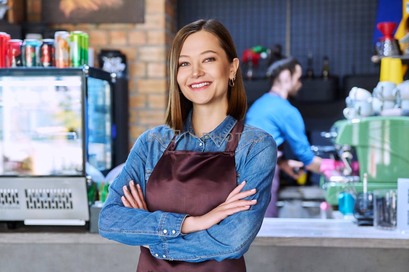 Young woman service worker in apron looking at camera in restaurant, coffee shop by VH-studio