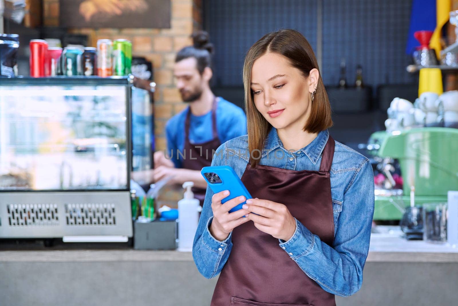 Confident successful young woman service worker in apron holding smartphone in restaurant cafeteria coffee shop pastry shop. Small business, staff, occupation, entrepreneur owner, work concept