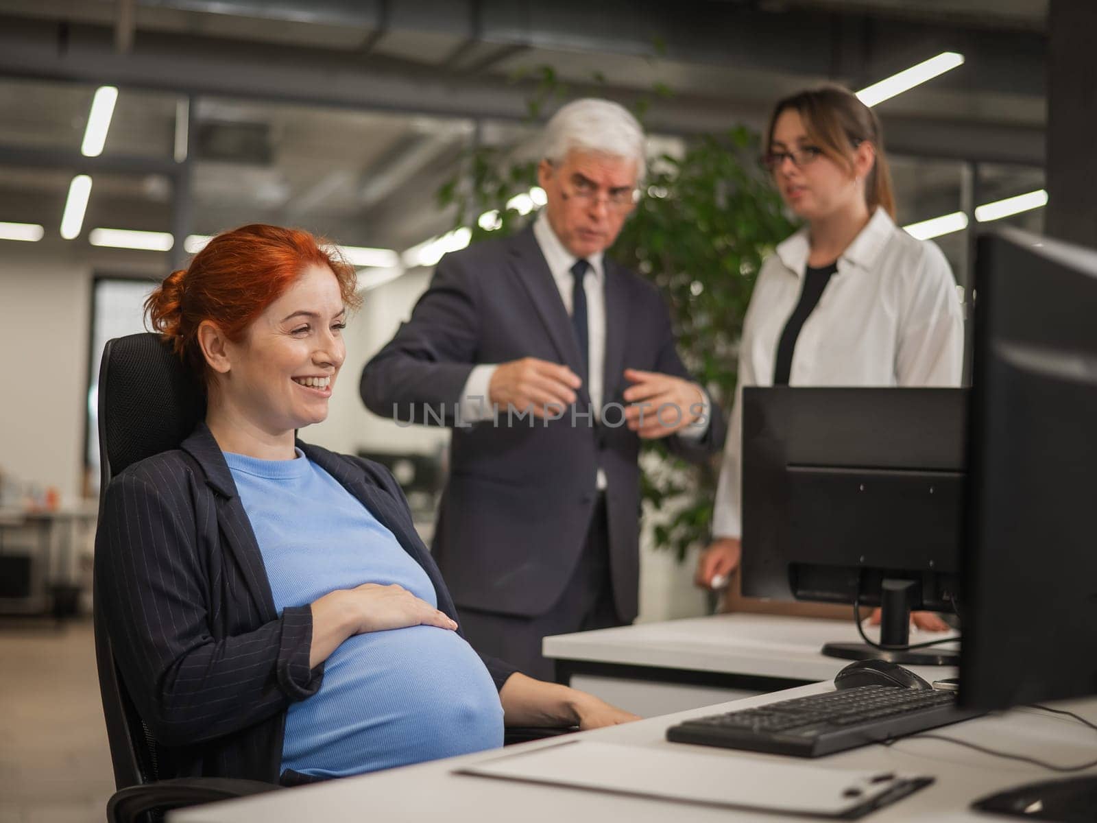 Happy Caucasian pregnant woman sitting at her desk, colleagues gossiping behind her back. by mrwed54