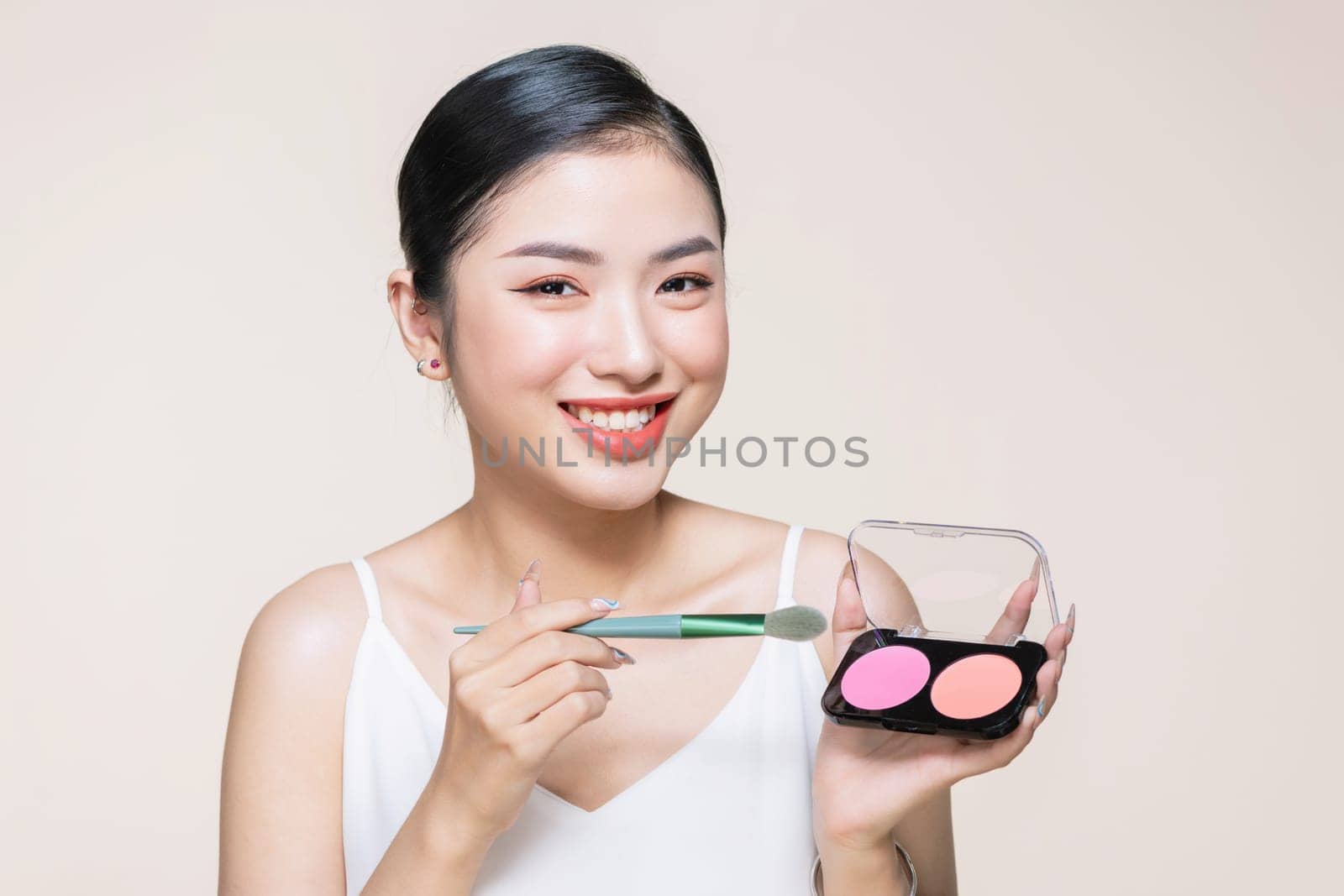 Young asian smiling woman holding blush palette with a brush isolated on a beige background