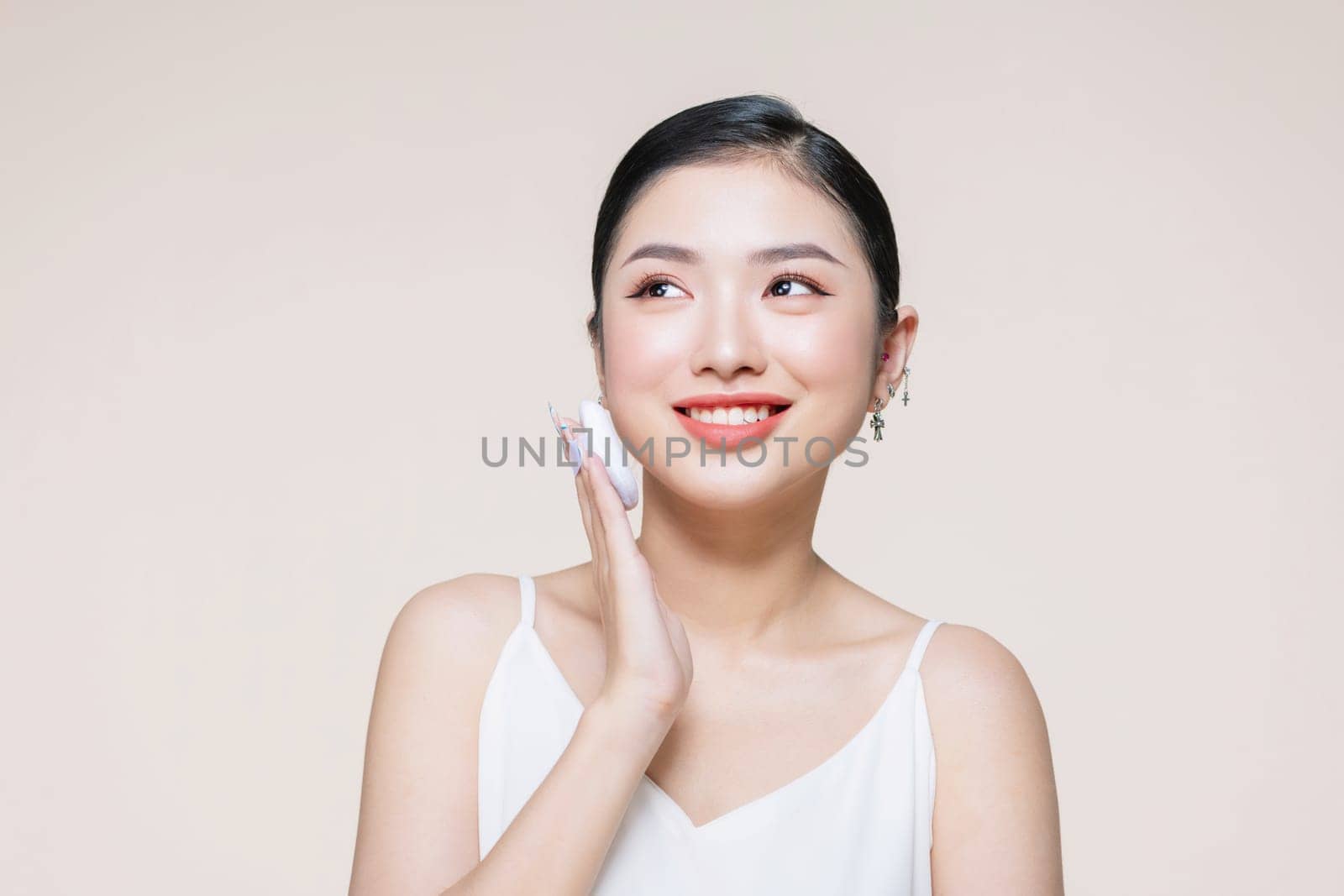 Young asian woman smile looking happy applying makeup with puff makeup on beige background.