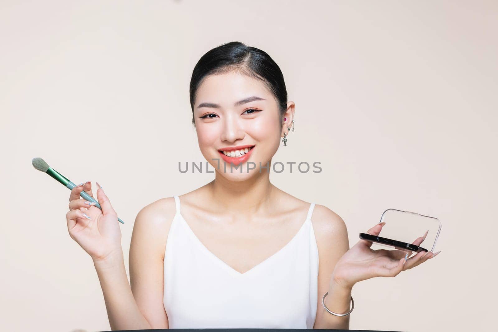 Young asian woman doing makeup with brush while standing on beige background