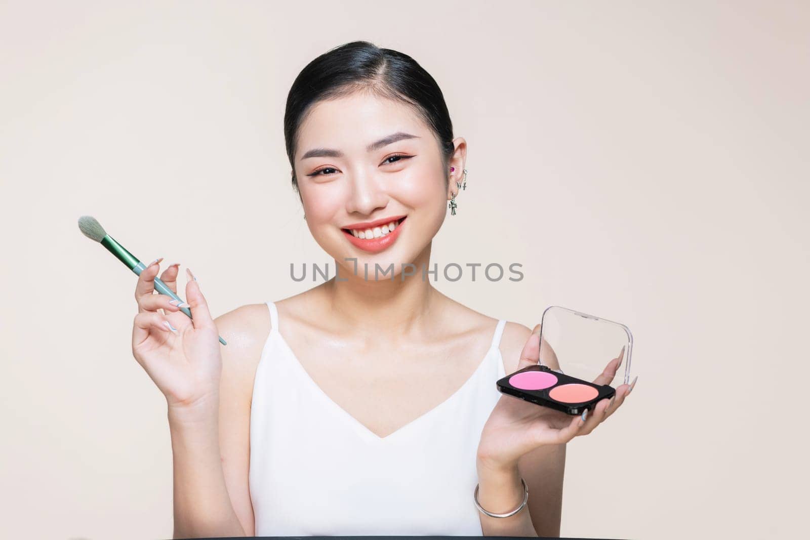 Young asian smiling woman holding blush palette with a brush isolated on a beige background