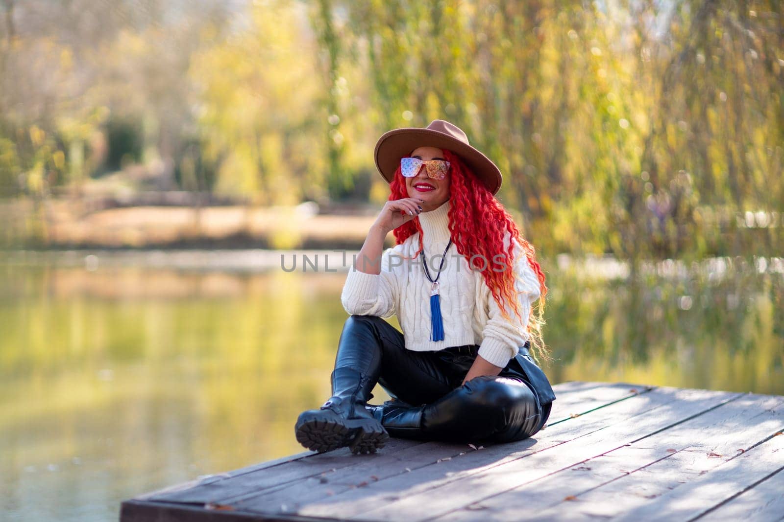 Autumn lake woman. In autumn, she sits by the pond on a wooden pier and admires nature with red hair and a hat. Tourism concept, weekend outside the city. by Matiunina