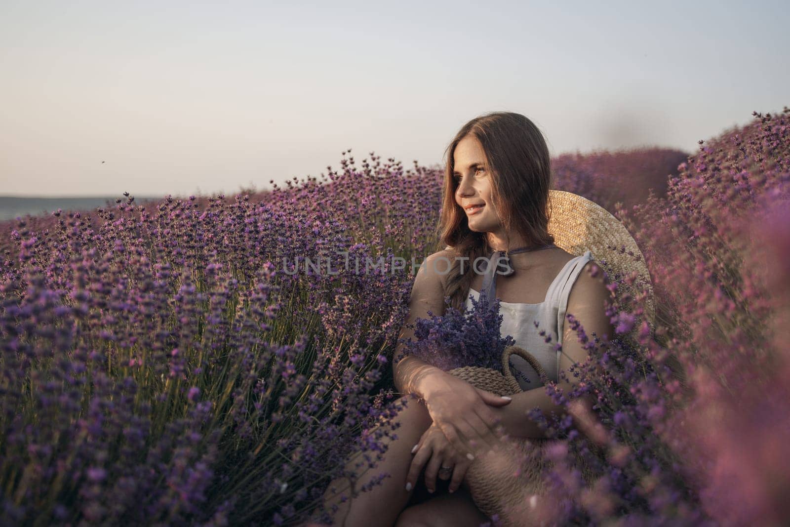 A woman is sitting in a field of lavender flowers. She is wearing a straw hat and holding a basket of flowers. The scene is peaceful and serene, with the woman enjoying the beauty of the flowers. by Matiunina