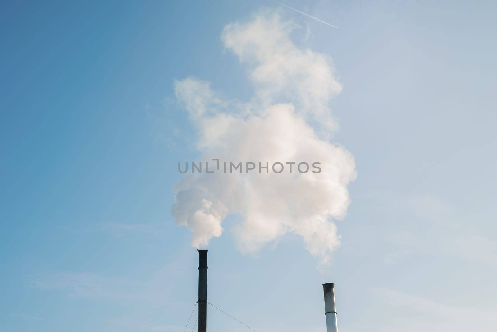 Two smoking factory chimneys and an airplane trail against a blue sky. Increase CO2 and greenhouse gas emissions.