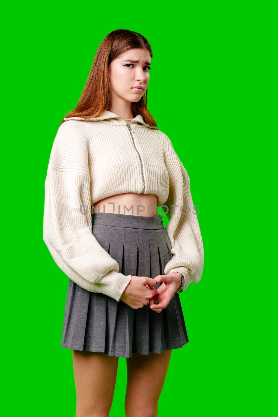 A young woman is standing with her hands gently clasped in front of her. She is wearing a white knitted cropped sweater and a pleated grey skirt. Her expression is contemplative, and she gazes off to the side. The solid green backdrop suggests a setup for a chroma key compositing.