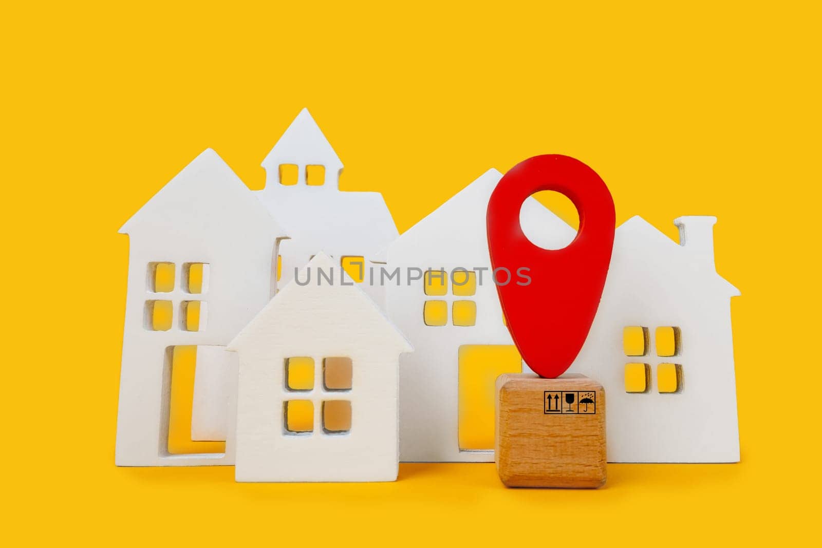 Cardboard boxes and a red position pin in city of white toy house on yellow background. Locating packages and goods.