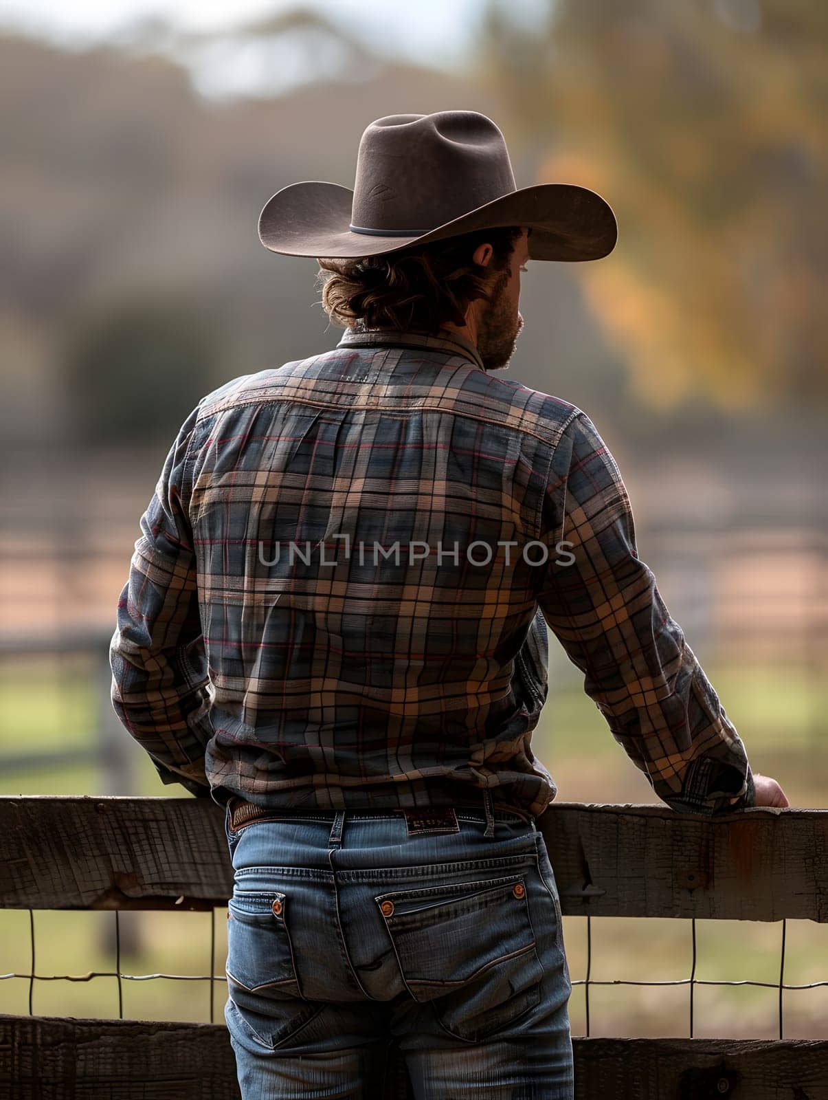 A man in jeans and a dress shirt is leaning on a wooden fence, wearing a cowboy hat and enjoying the landscape in his fedora hat