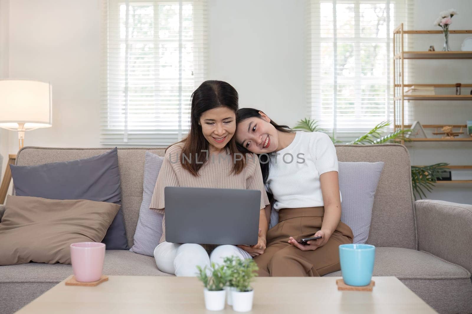 Mother and teenage daughter are looking at social media on laptop together on the sofa in the living room. by wichayada