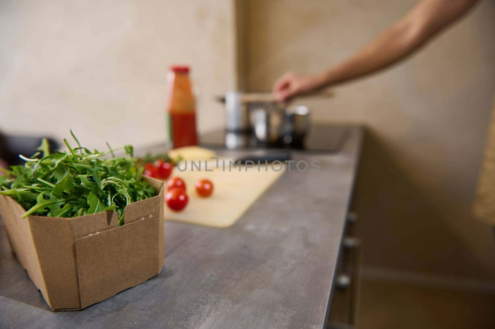 Healthy fresh organic arugula herbs in cardboard box, ingredients and blurred stainless steel pan on electric stove in the home kitchen interior. Healthy ingredients for Mediterranean lunch. Culinary.