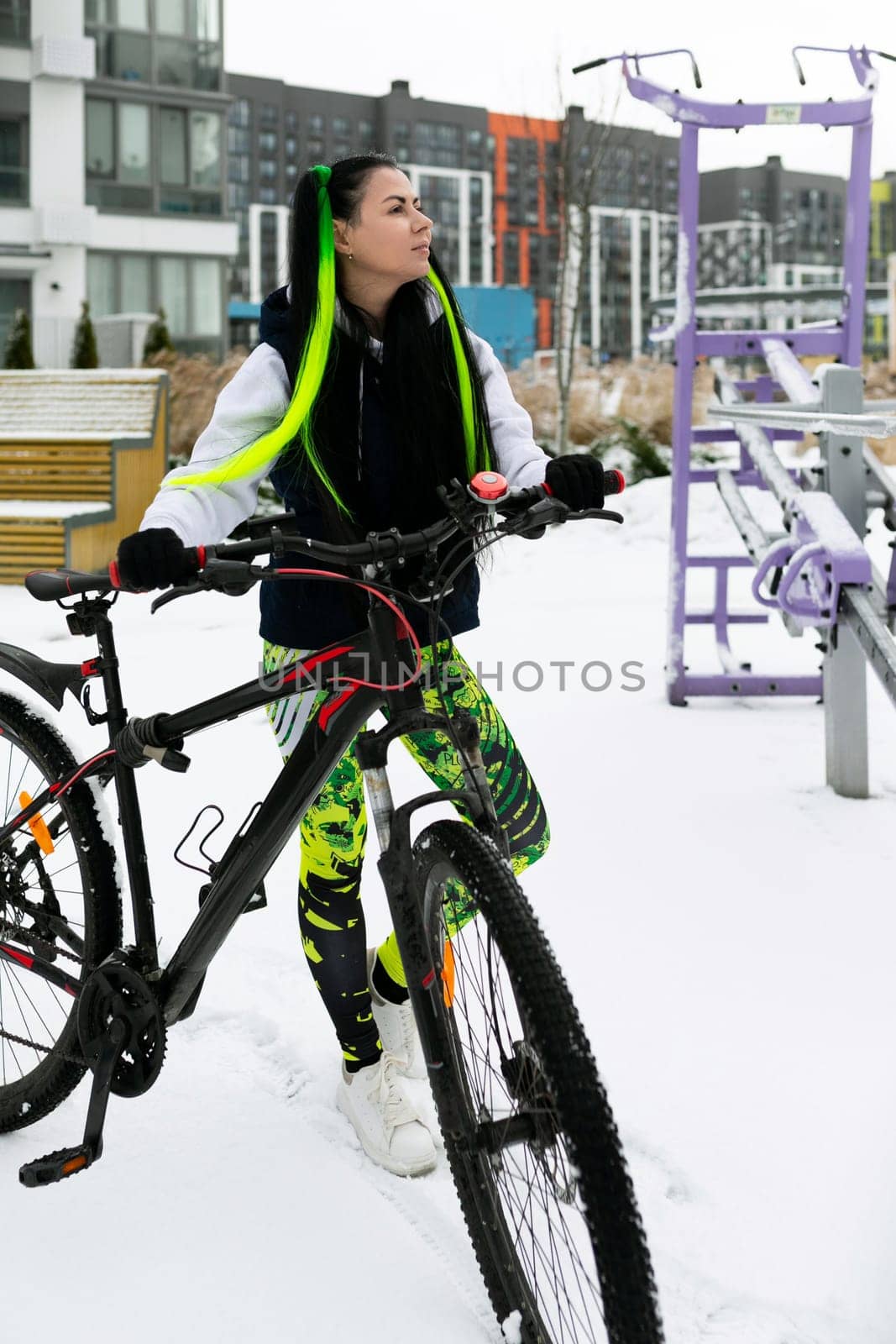 A woman wearing winter clothes is standing next to a parked bicycle in a snow-covered environment. She appears to be observing her surroundings while the bike is covered in a thin layer of snow.