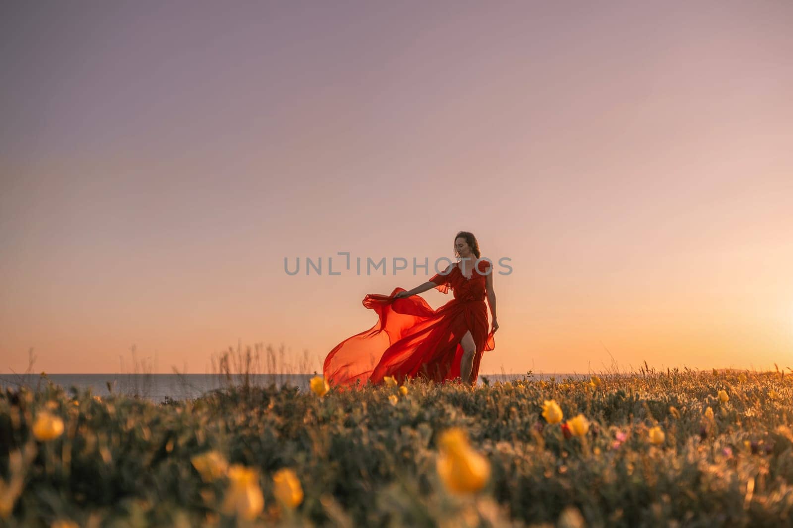 woman red dress is standing on a grassy hill overlooking the ocean. The sky is a beautiful mix of orange and pink hues, creating a serene and romantic atmosphere