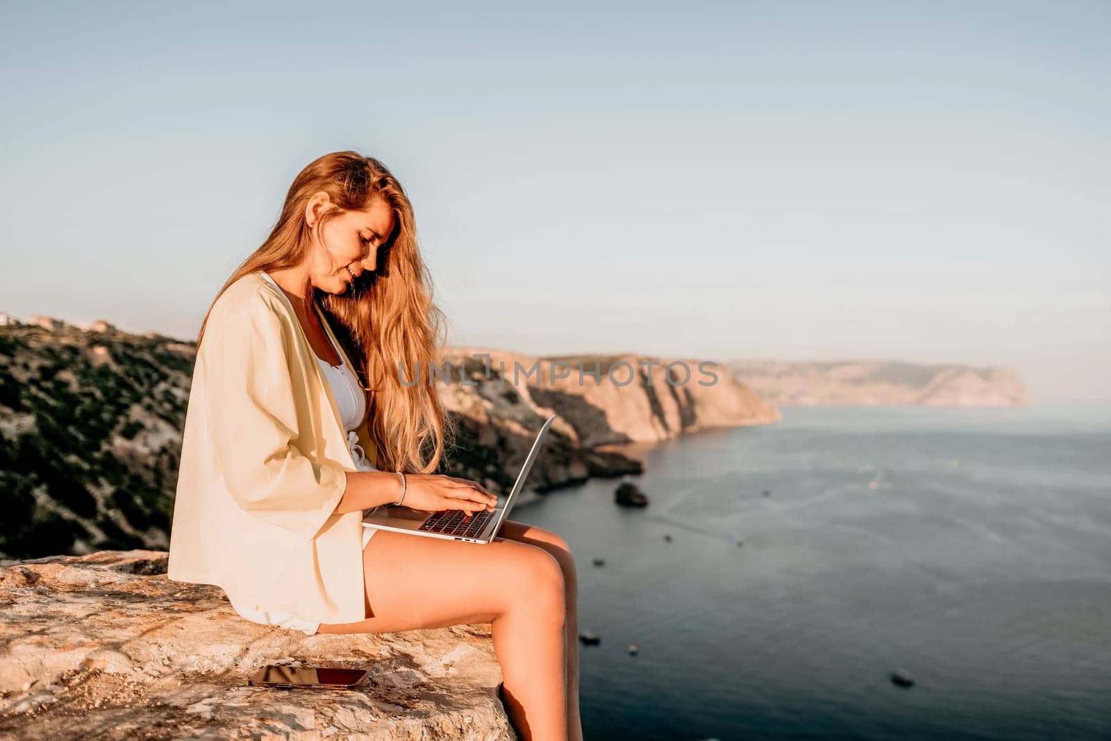 Successful business woman in yellow hat working on laptop by the sea. Pretty lady typing on computer at summer day outdoors. Freelance, travel and holidays concept.