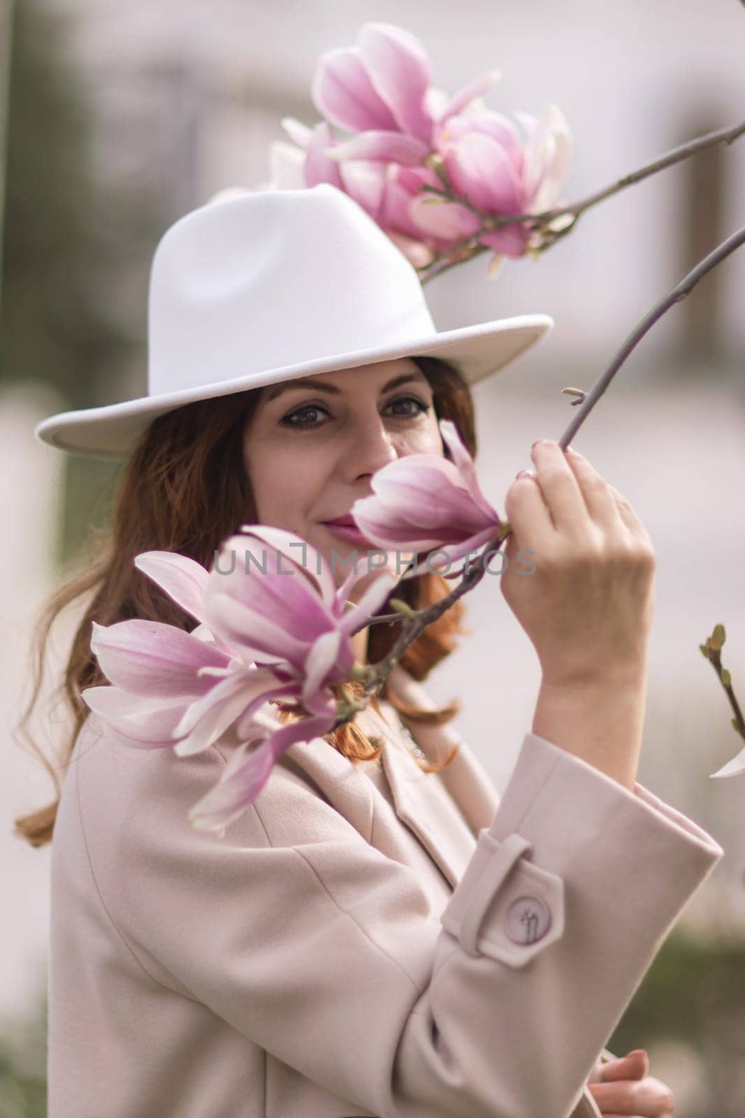 Woman magnolia flowers, surrounded by blossoming trees, hair down, white hat, wearing a light coat. Captured during spring, showcasing natural beauty and seasonal change