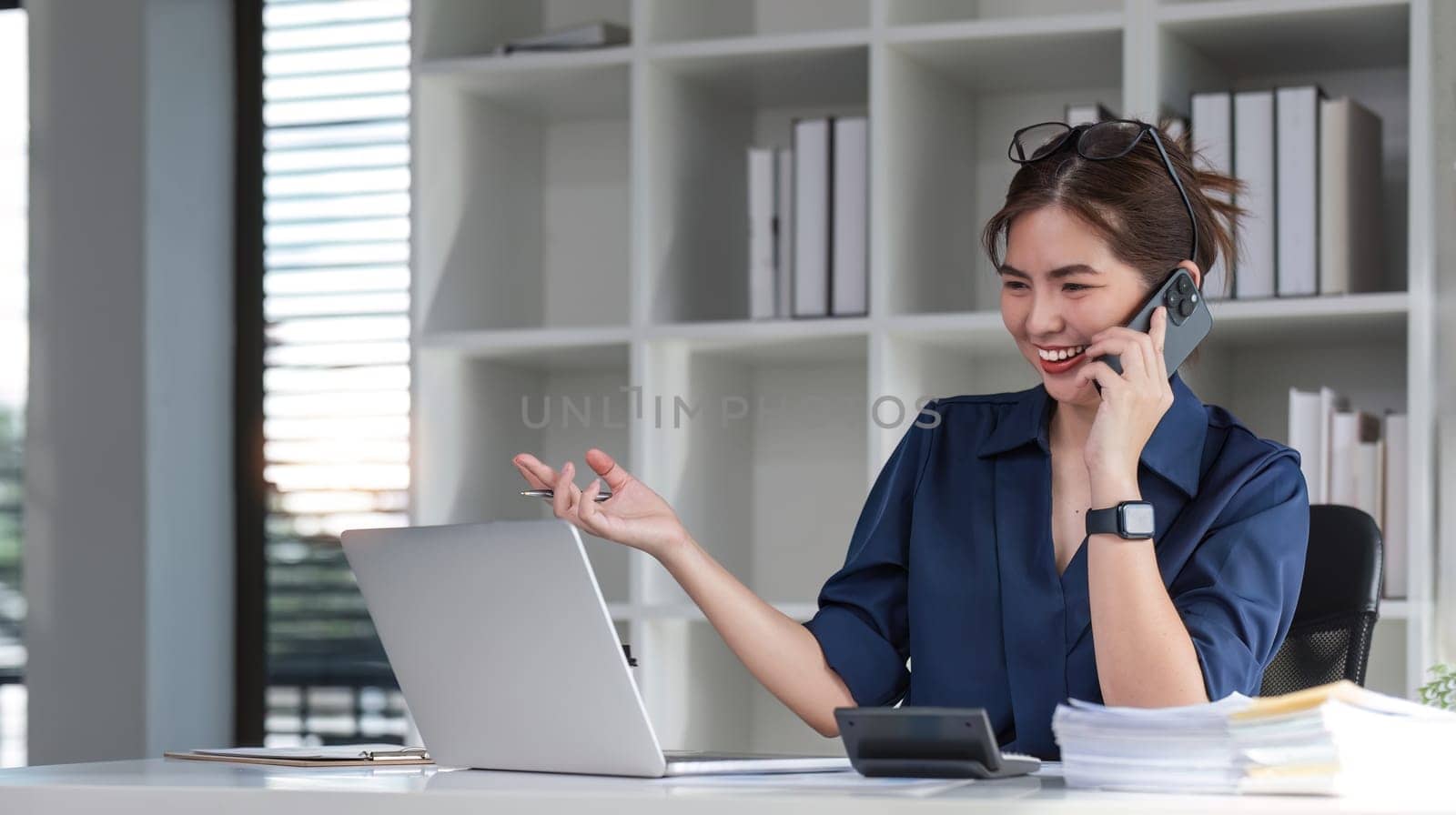 Young businesswoman using the phone talking working on starting a business and calculating finances with a calculator. by wichayada