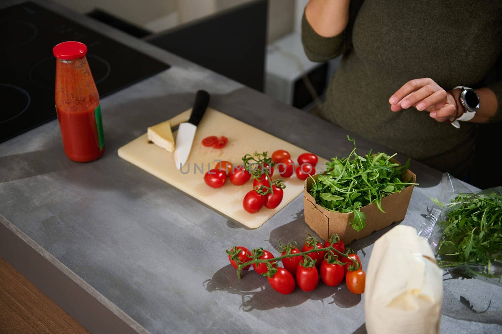 Close-up unknown woman standing at kitchen counter with a fresh bunch of tomato cherry, arugula leaves and Italian pasta in paper packet. Healthy eating, culinary, diet concept. Copy advertising space