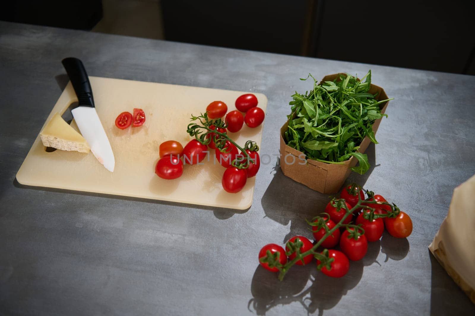 Top view of fresh organic ingredients for a healthy salad. Eco-friendly cardboard box with arugula leaves, a bunch of red ripe cherry tomatoes and a slice of parmesan cheese on a cutting board. by artgf