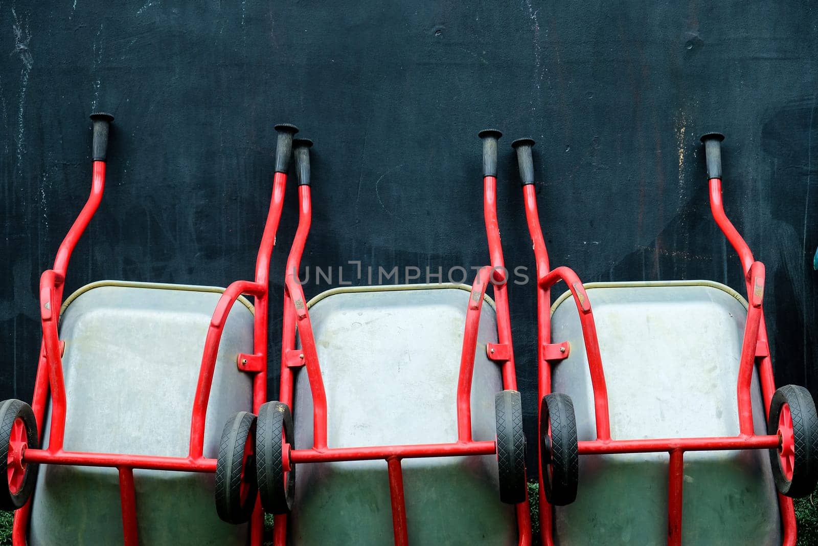 Array of red wheelbarrows lined up against a dark textured wall by ponsulak