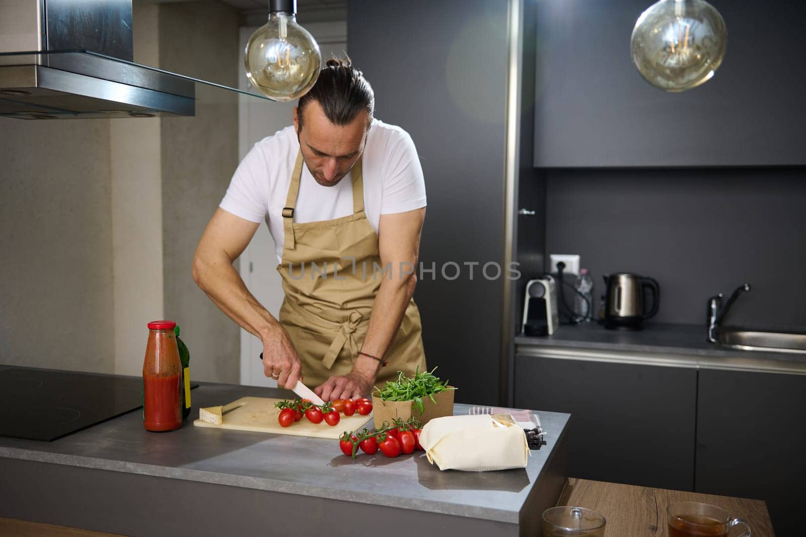 Handsome young man in beige chef's apron, cooking healthy vegetarian salad in the modern minimalist home kitchen, standing at kitchen island and chopping fresh ripe cherry tomatoes. by artgf