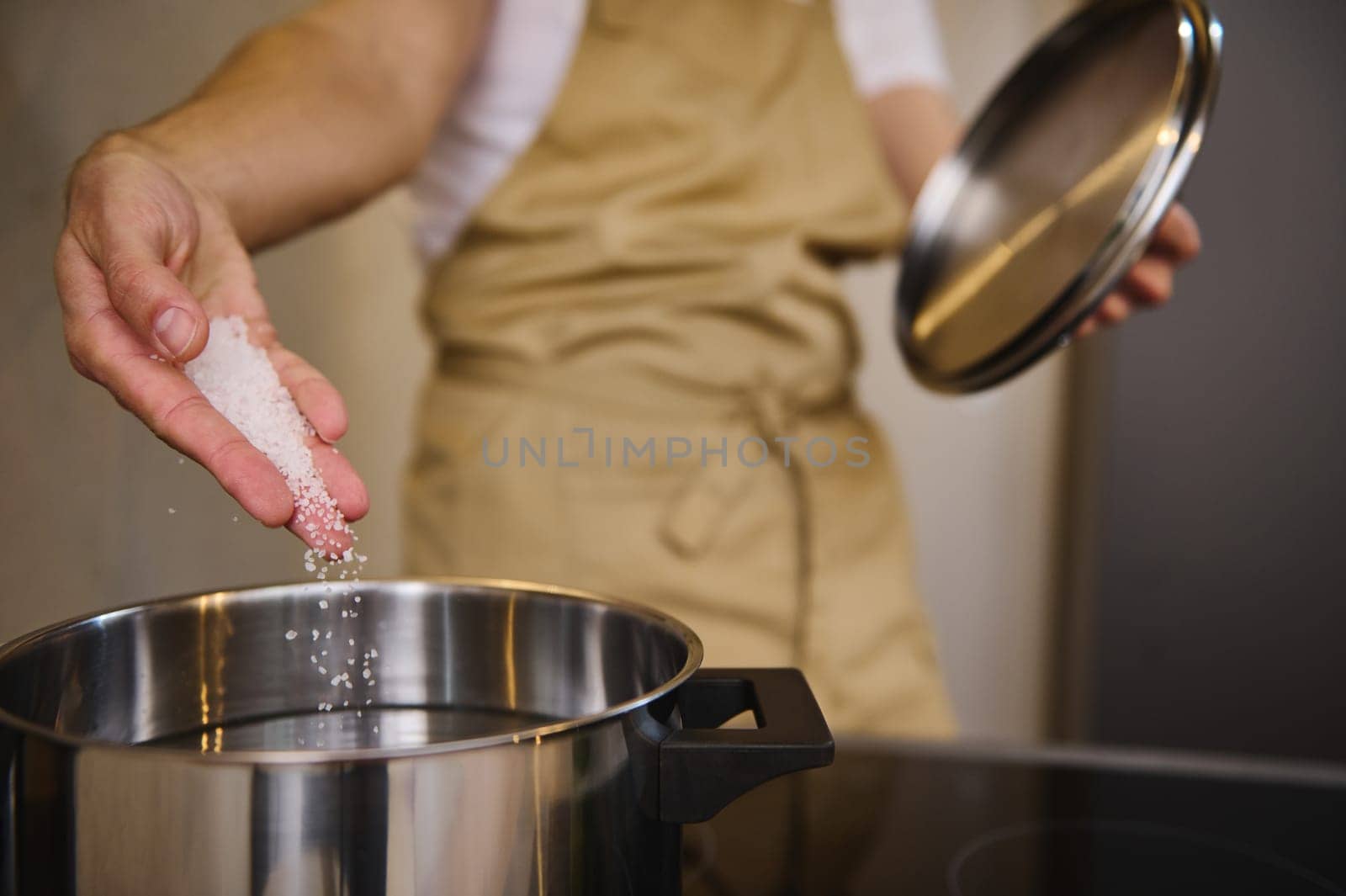 Close-up of unrecognizable chef adding salt into a boiling water in the stainless steel saucepan, standing at an electric stove, seasoning the boiling water for spaghetti or other meal. Food concept