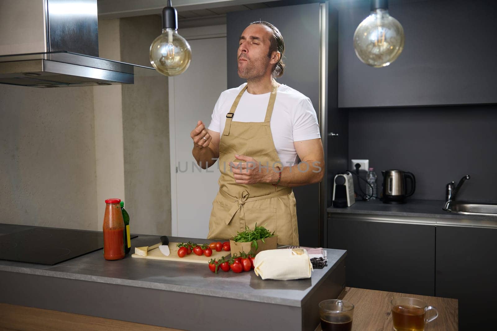 Handsome Hispanic young man tasting ingredients while cooking dinner at home kitchen, standing at kitchen table with ingredients, expressing satisfaction of the taste of veggies. People. Healthy food.