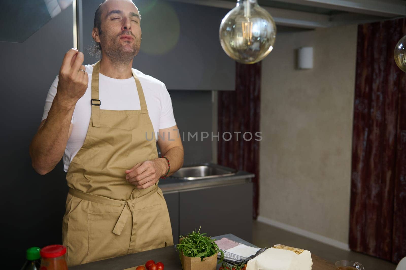 Young adult Caucasian male chef tasting ingredients, kissing fingers and making delicious finger gesture while cooking dinner in the home kitchen. People. Cooking at home. Culinary. Domestic life