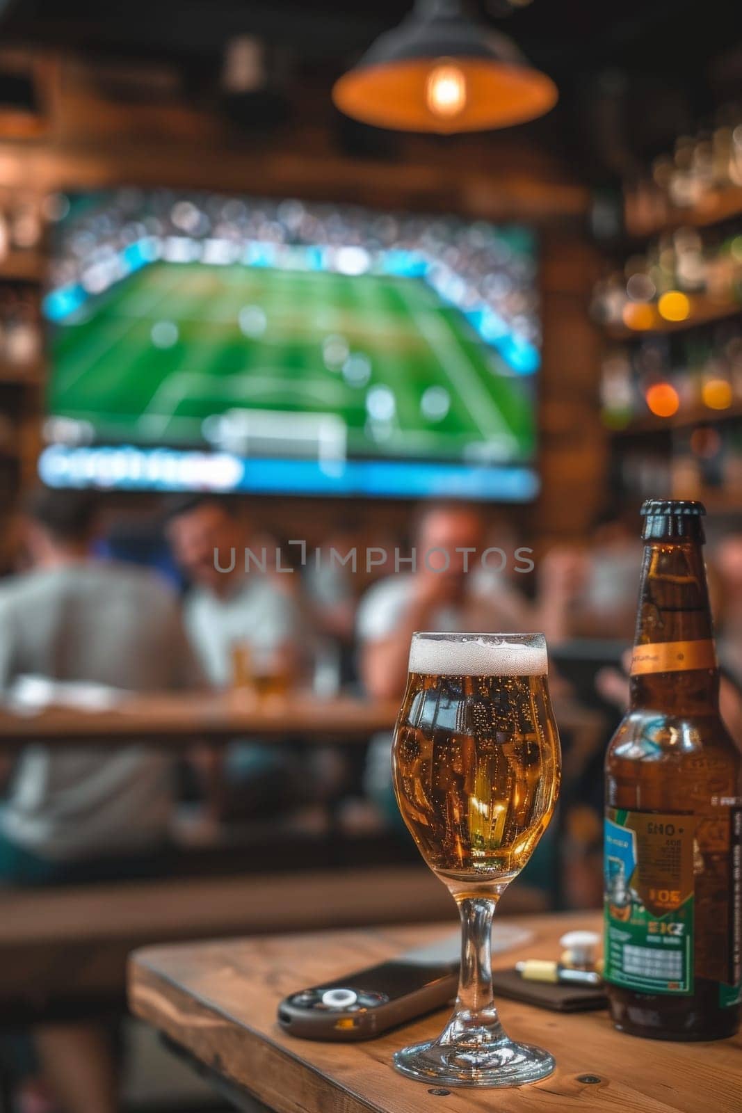 A glass of beer sits on a table next to a bottle of beer. The scene is set in a bar or pub, with a TV in the background showing a soccer game. The atmosphere is lively and social