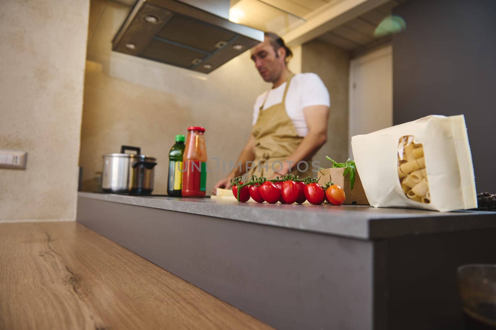 Selective focus on a bunch of red ripe organic tomato cherry and a paper packet with pasta, against male chef standing at induction stove, adjusting program, cooking dinner in modern kitchen interior
