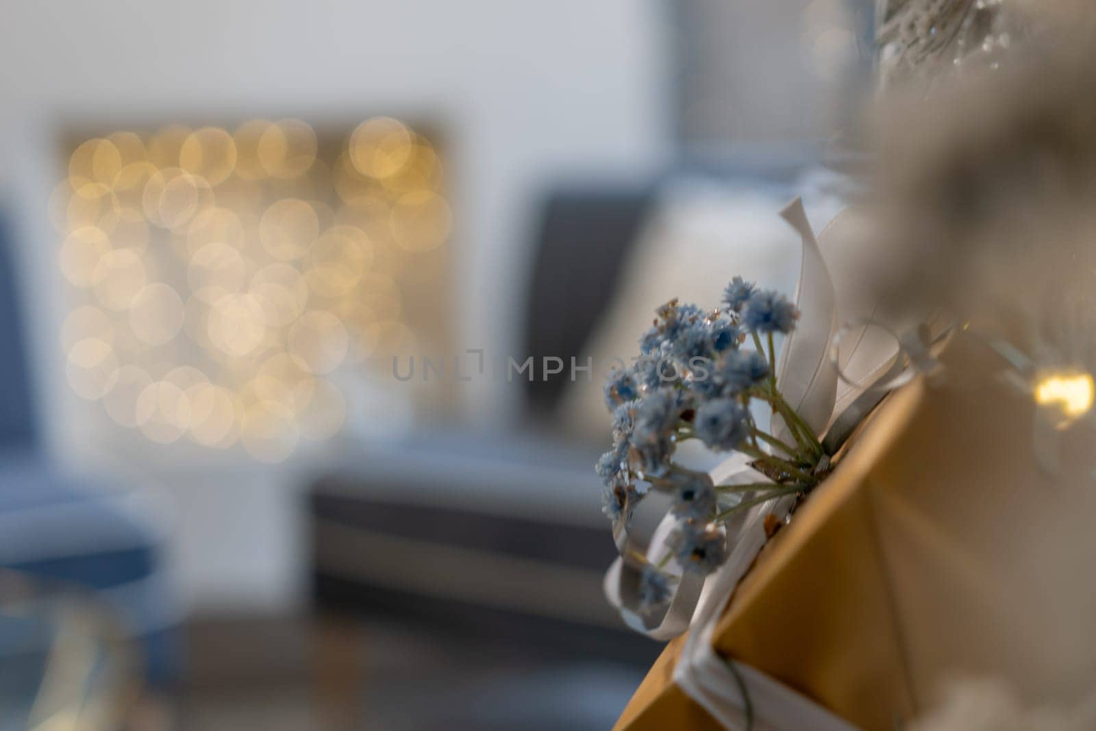 A bouquet of flowers is sitting on top of a gold box. The flowers are blue and white, and the box is wrapped in gold paper. The scene is set in a living room, with a couch. by Matiunina