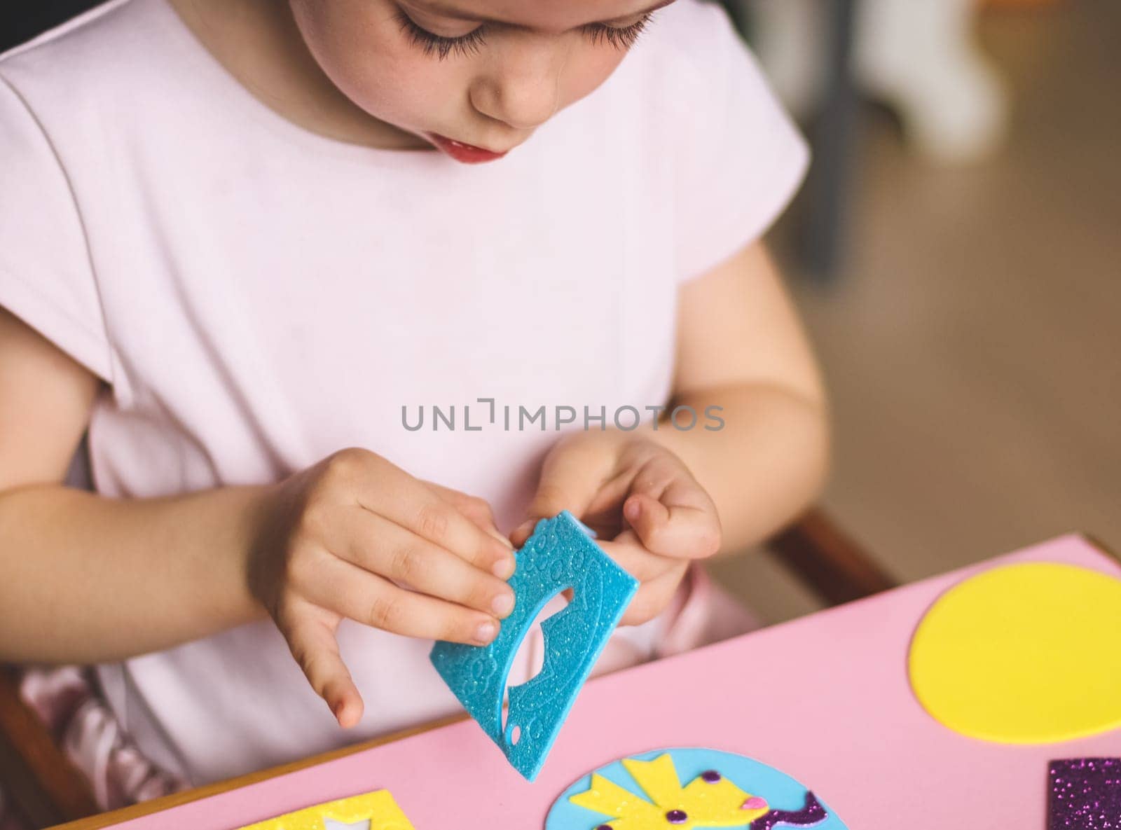 A little caucasian girl enthusiastically peels off with her fingers a blue circle felt sticker for an Easter egg with a yellow bow, sitting at a children's table with a set of crafts on a pink background with depth of field, close-up side view. Concept of crafts, diy, needlework, artisanal, children art, easter preparation, children creative.