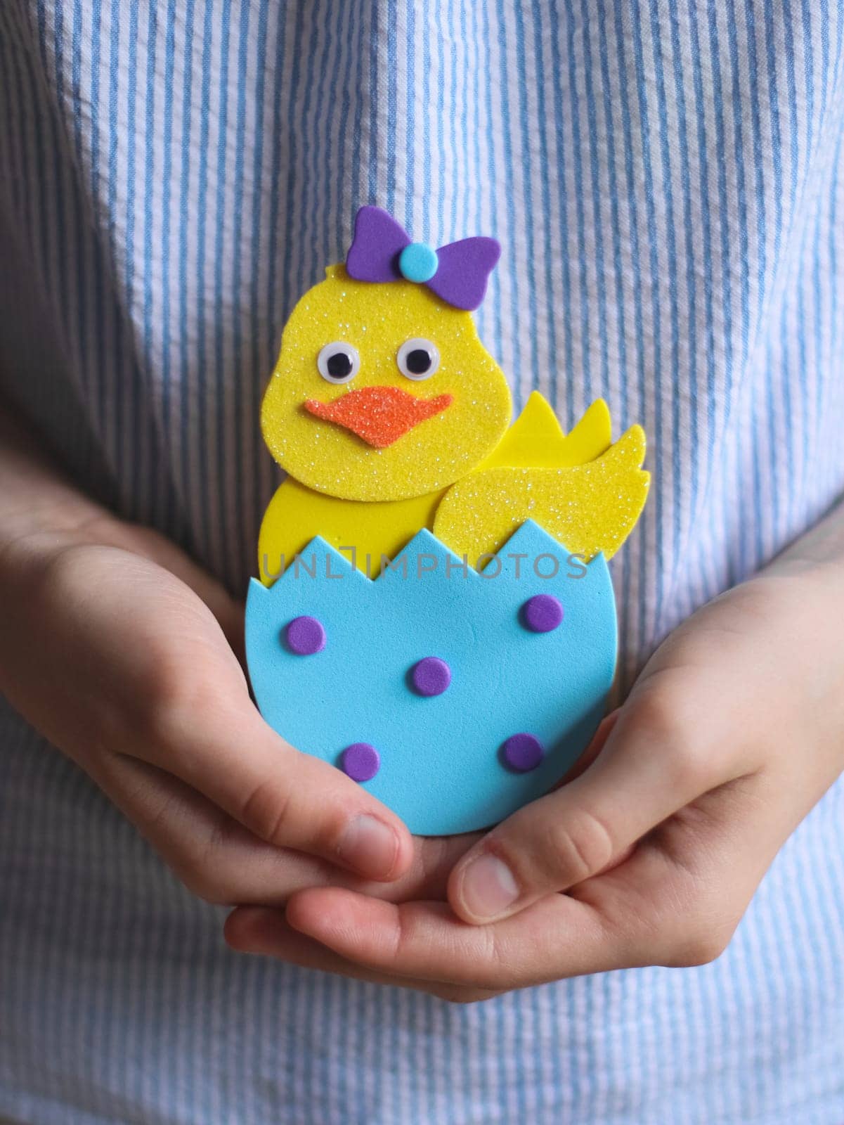 Hands of Caucasian teenage girl showing end result craft of yellow easter felt chicken with blue shell, orange beak, eyes, lilac bow and circles, holding in palms at belly level with blue striped blouse with depth of field, closeup side view. Concept of crafts, diy, needlework, artisanal, children art, easter preparation, children creative.