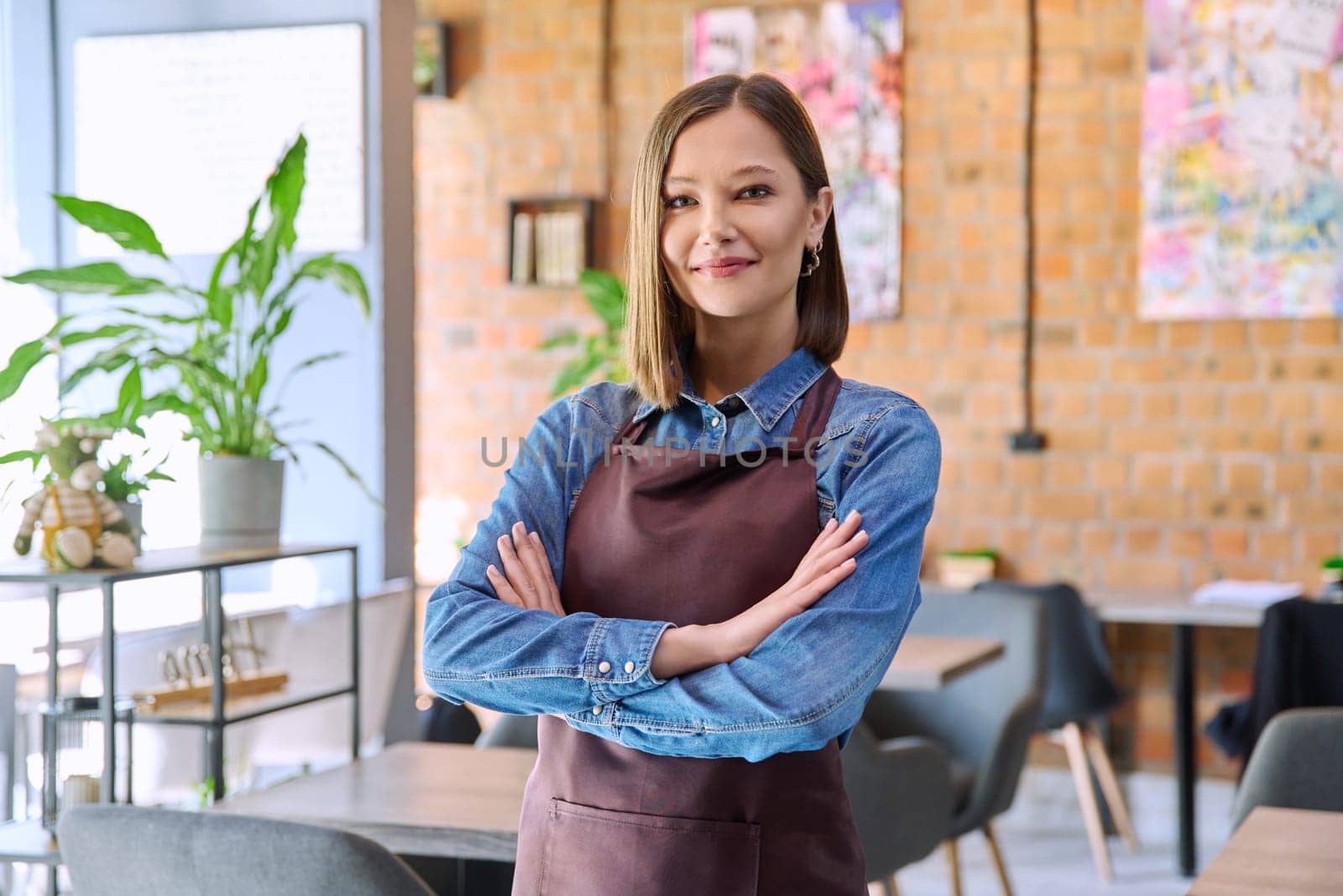 Confident successful young woman service worker owner in apron with crossed arms looking at camera in restaurant cafeteria coffee pastry shop interior. Small business staff occupation entrepreneur work