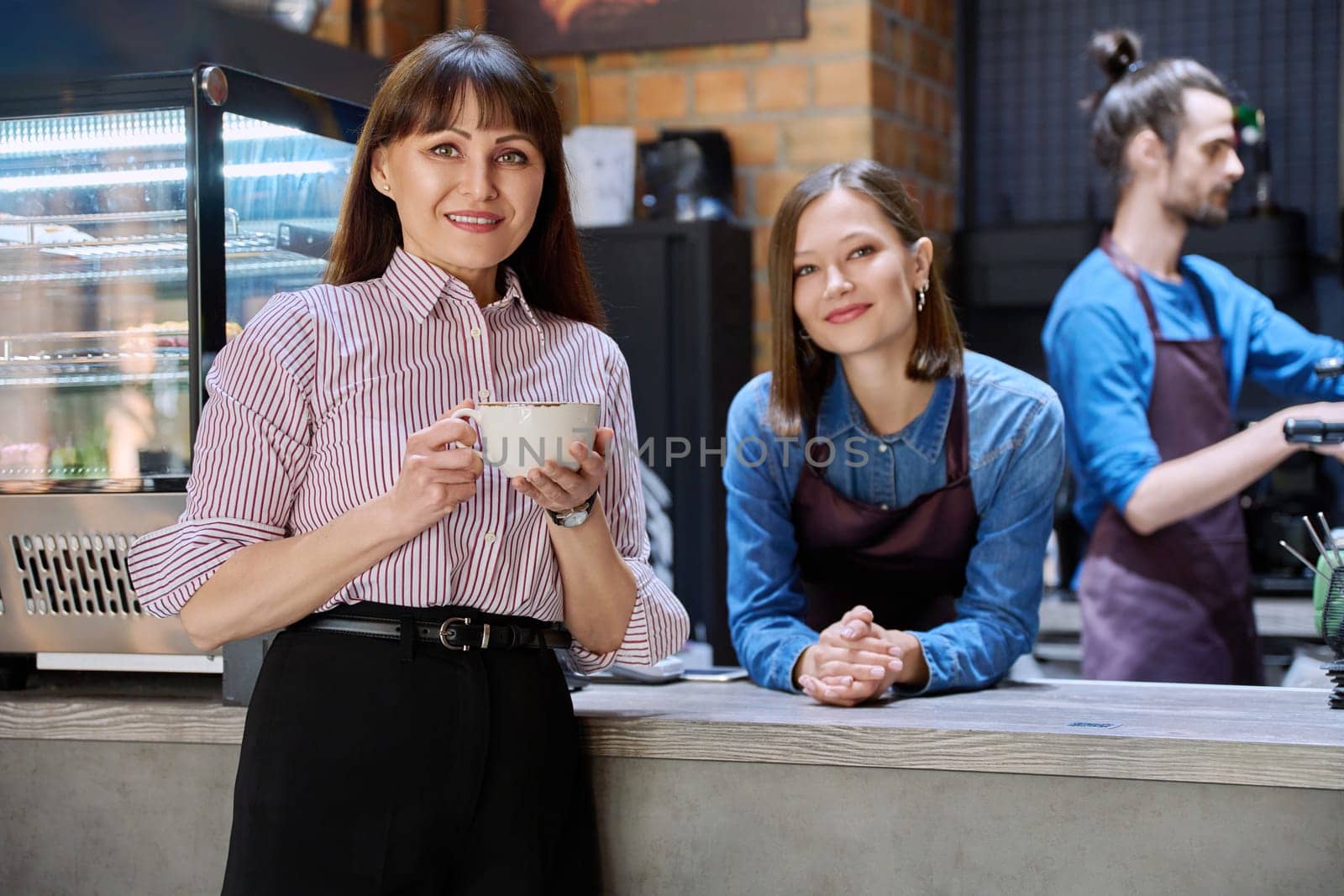 Woman customer of coffee shop near counter with cup of coffee looking at camera, restaurant workers owners at workplace. Colleagues partners in food service work, entrepreneurship, small business