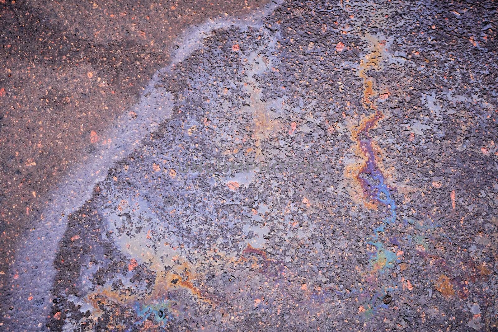 Oil or gasoline leaking from under a car in the rain on the asphalt in a parking lot. Detail of an oil stain on the asphalt of a wet road. by AliaksandrFilimonau