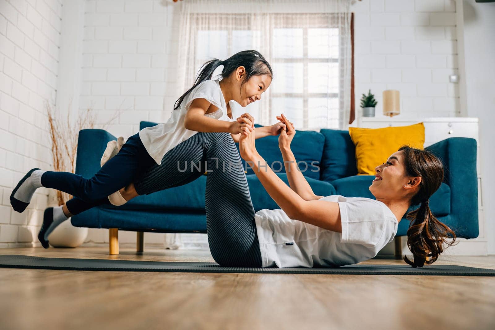 In a heartwarming family yoga session at home a mother practices the little bird posture holding her daughter's legs and fostering trust bonding and happiness.