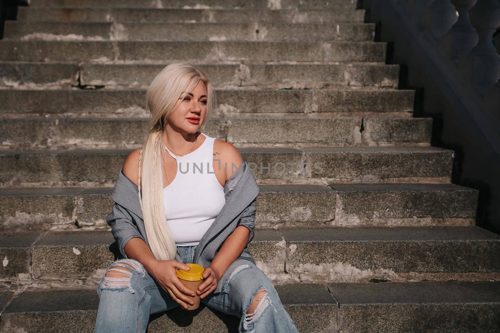 A blonde woman sits on a set of stairs, holding a cup. She is wearing a gray jacket and blue jeans. by Matiunina